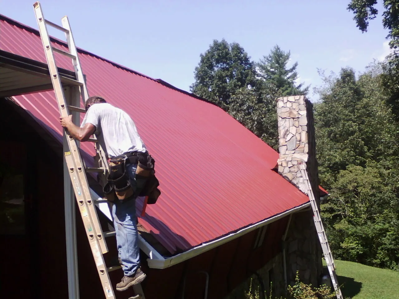 Roofer on a ladder beside a red metal roof and stone chimney — Soffit & Fascia Repair by Roofing Brothers in Westlake